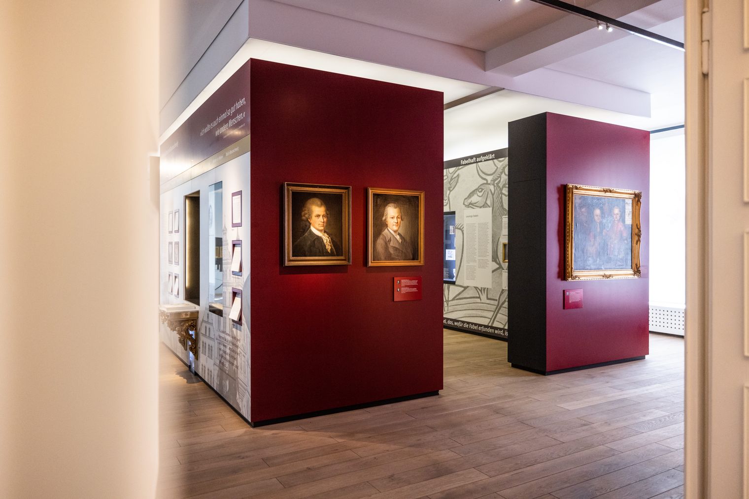 Innenansicht des Lessingmuseums Kamenz mit zwei Porträts an einer roten Wand und einem weiteren Gemälde an einer angrenzenden Wand. / Interior view of Lessingmuseum Kamenz showing two portraits on a red wall and another painting on an adjacent wall.