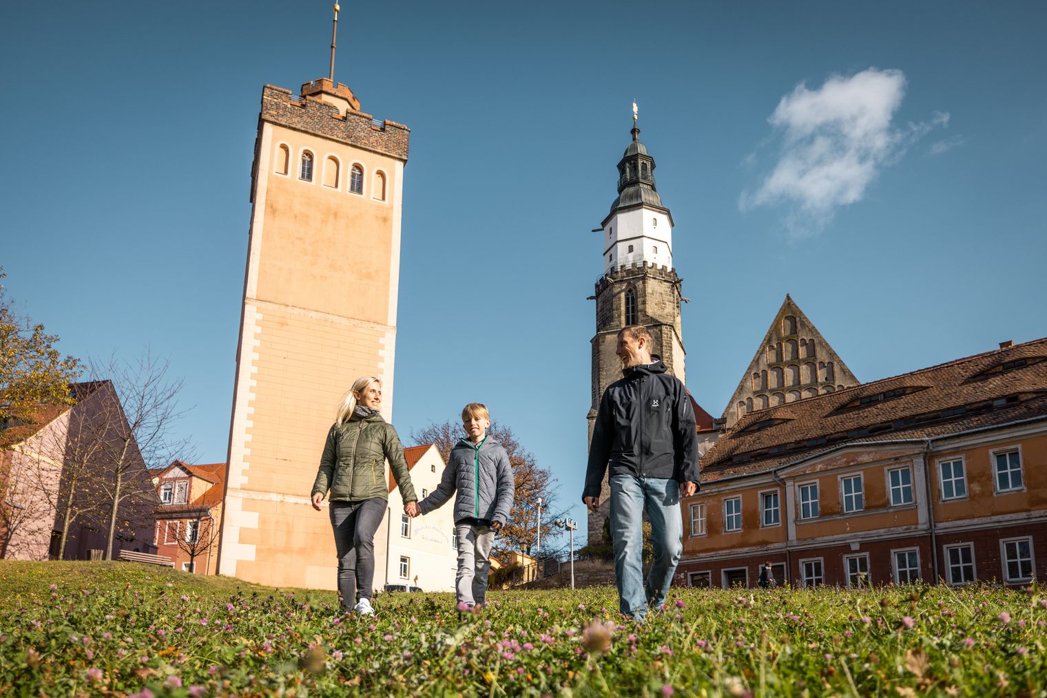Drei Personen gehen auf einer Wiese vor dem Roten Turm und der Kirche St. Marien in Kamenz bei klarem Himmel. / Three people walking on grass in front of the Roter Turm and St. Marien church in Kamenz under clear sky.