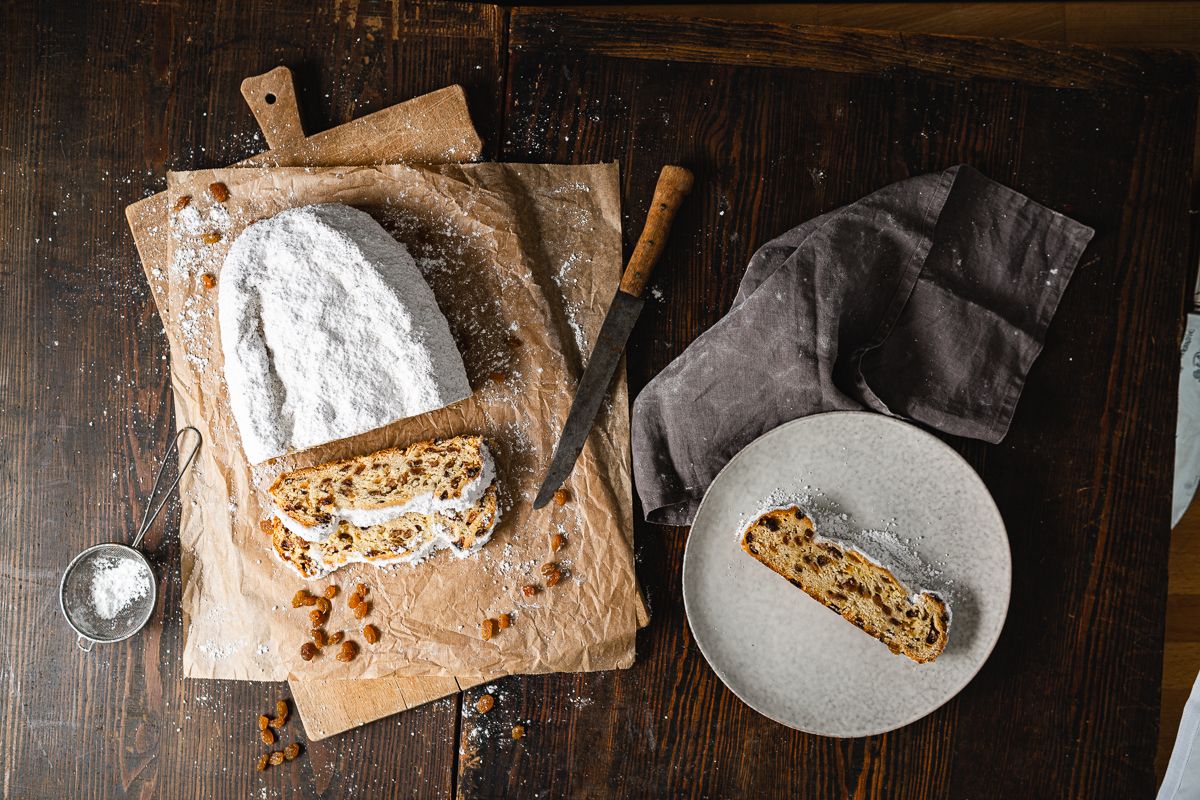 Ein Dresdner Christstollen liegt auf einem Holzbrett. Der Dresdner Stollen wurde mit einem Messer angeschnitten. Auf einem Teller befindet sich eine Scheibe des Dresdner Christstollens. 