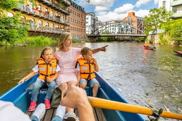 Eine Mutter und zwei Kinder sitzen an der Spitze eines Ruderbootes. Sie fahren über den Kanal in Leipzig.